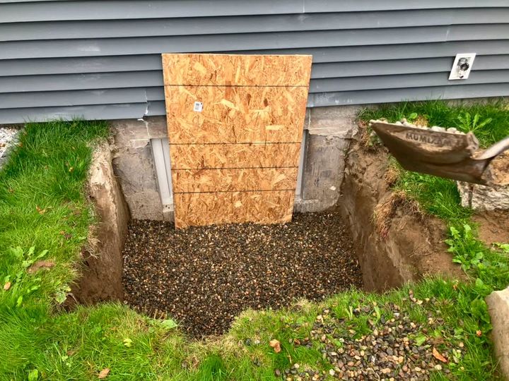 Before: Excavated window well pit with drainage rock and plywood over the opening
